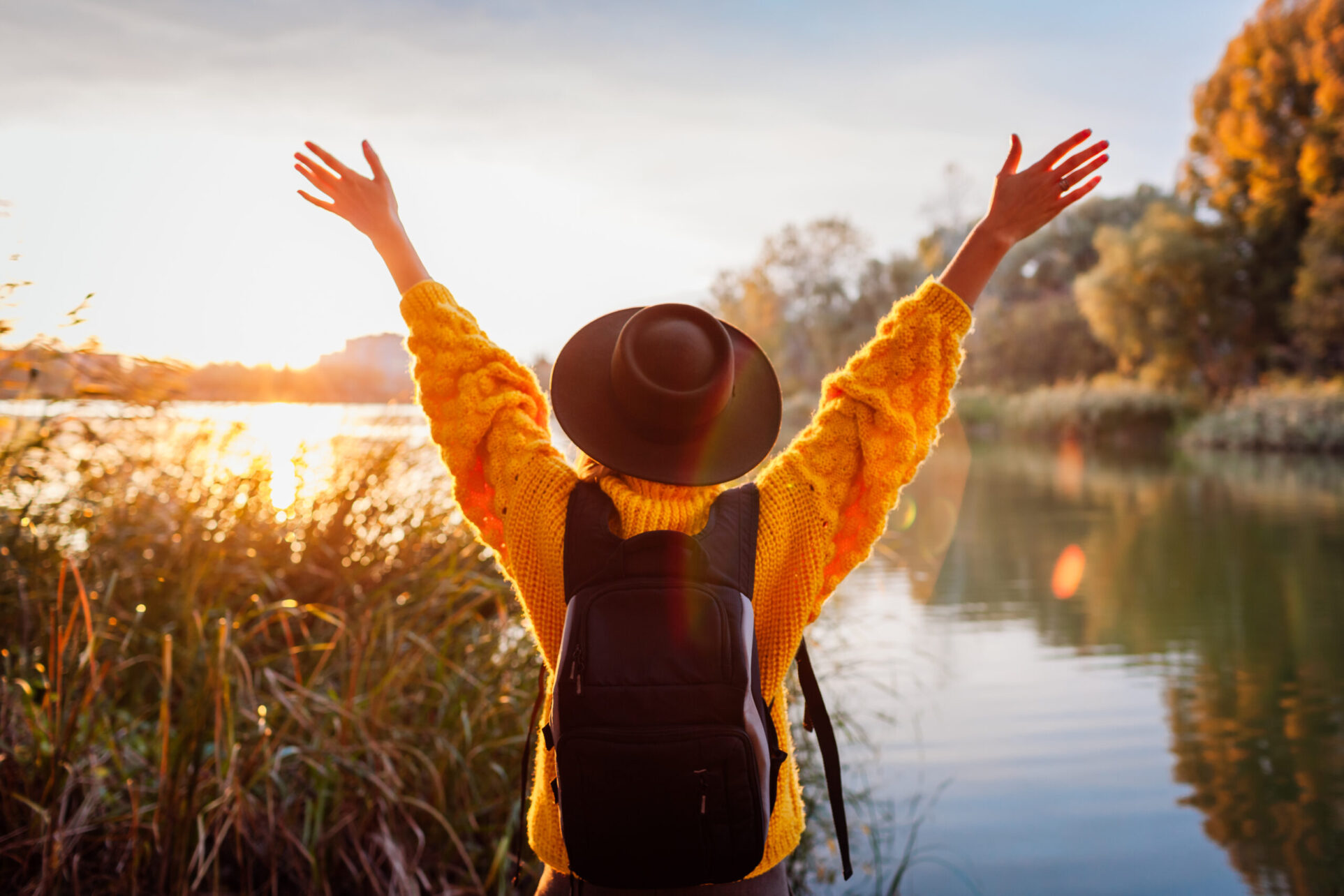 Traveler with backpack relaxing by autumn river at sunset. Young woman raised arms feeling free and happy